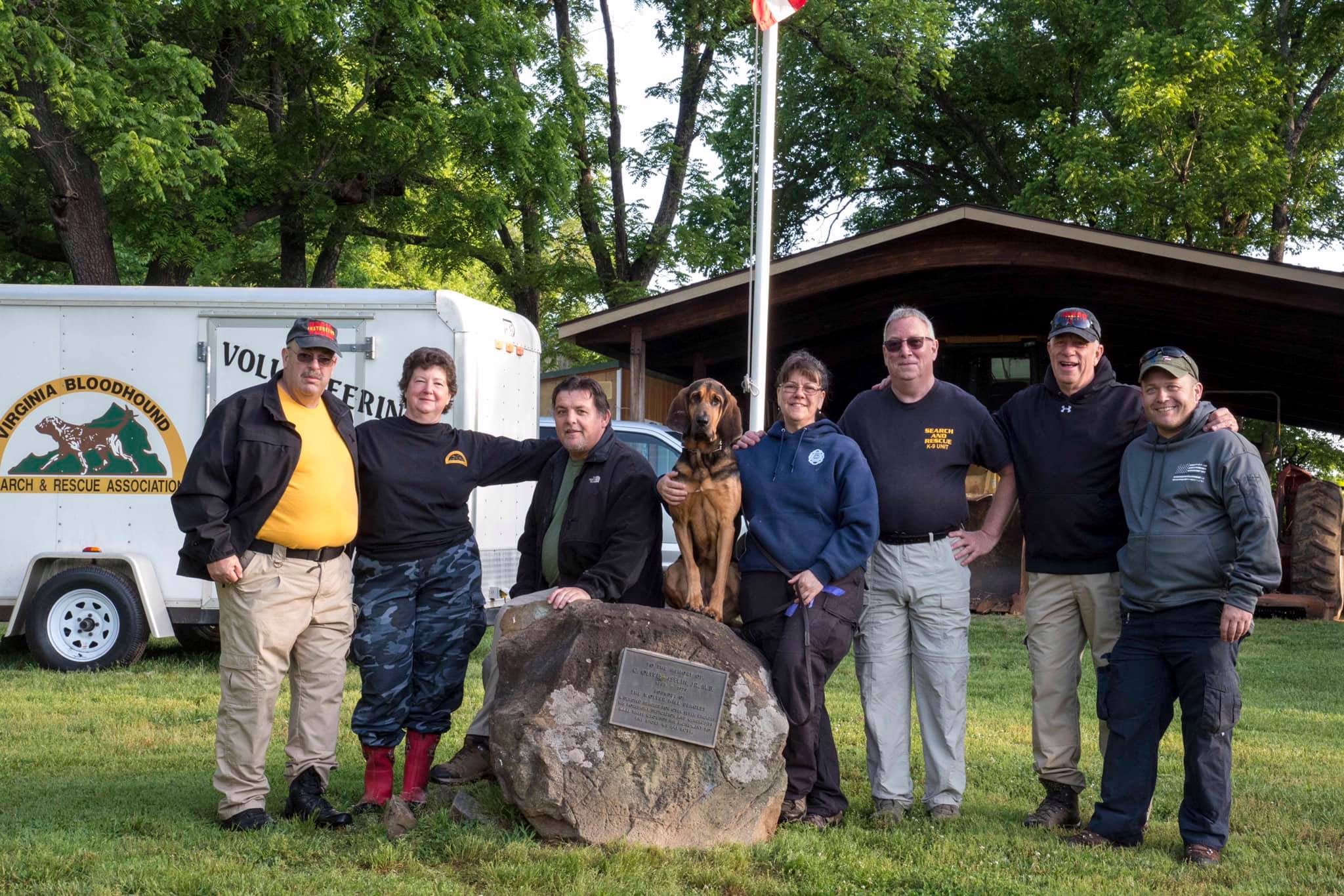 Search Dogs Northeast Search and Rescue Team stands on grass behind a small boulder with a bloodhound sitting atop it. There is a trailer and small building behind them with trees in the far background.